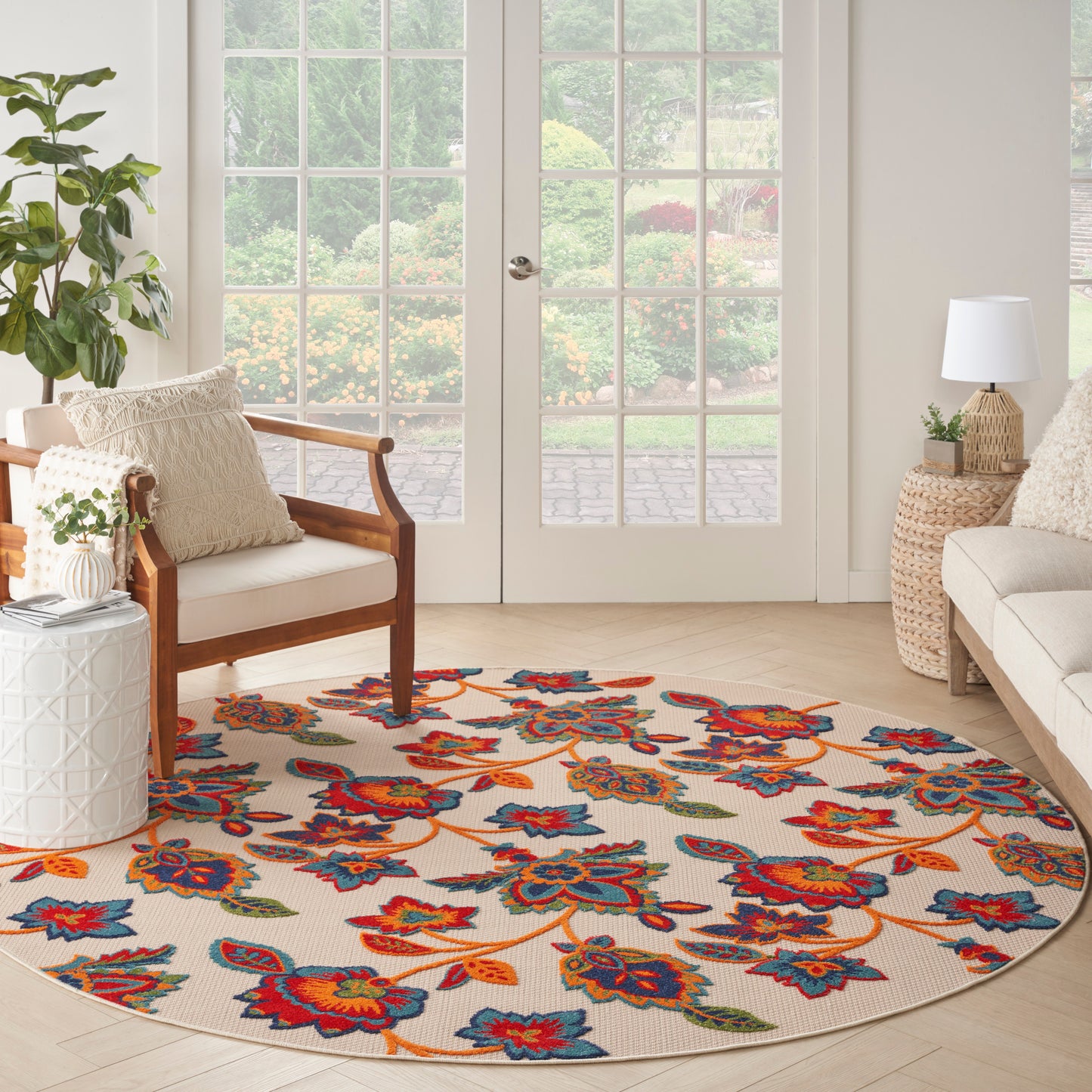 Round beige rug with bold multicolor floral pattern in a sunlit living room with wood-frame chairs and glass-paneled doors.