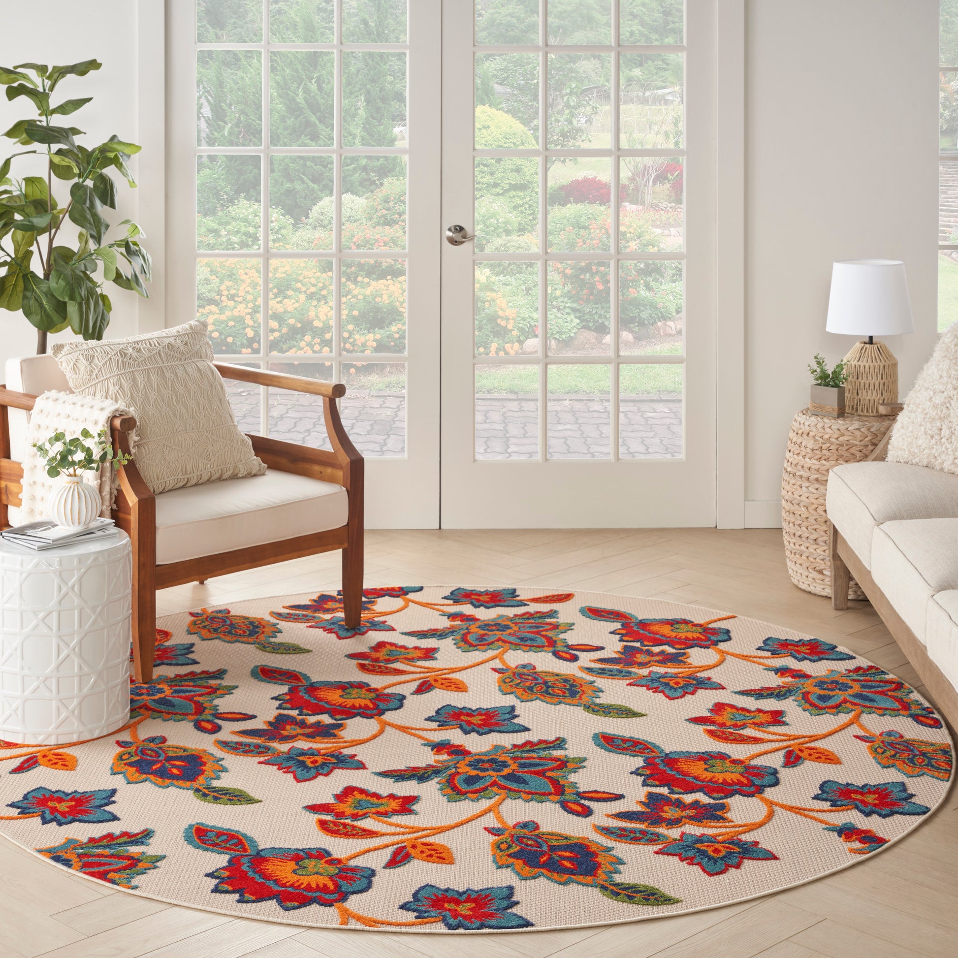 Round beige rug with bold multicolor floral pattern in a sunlit living room with wood-frame chairs and glass-paneled doors.