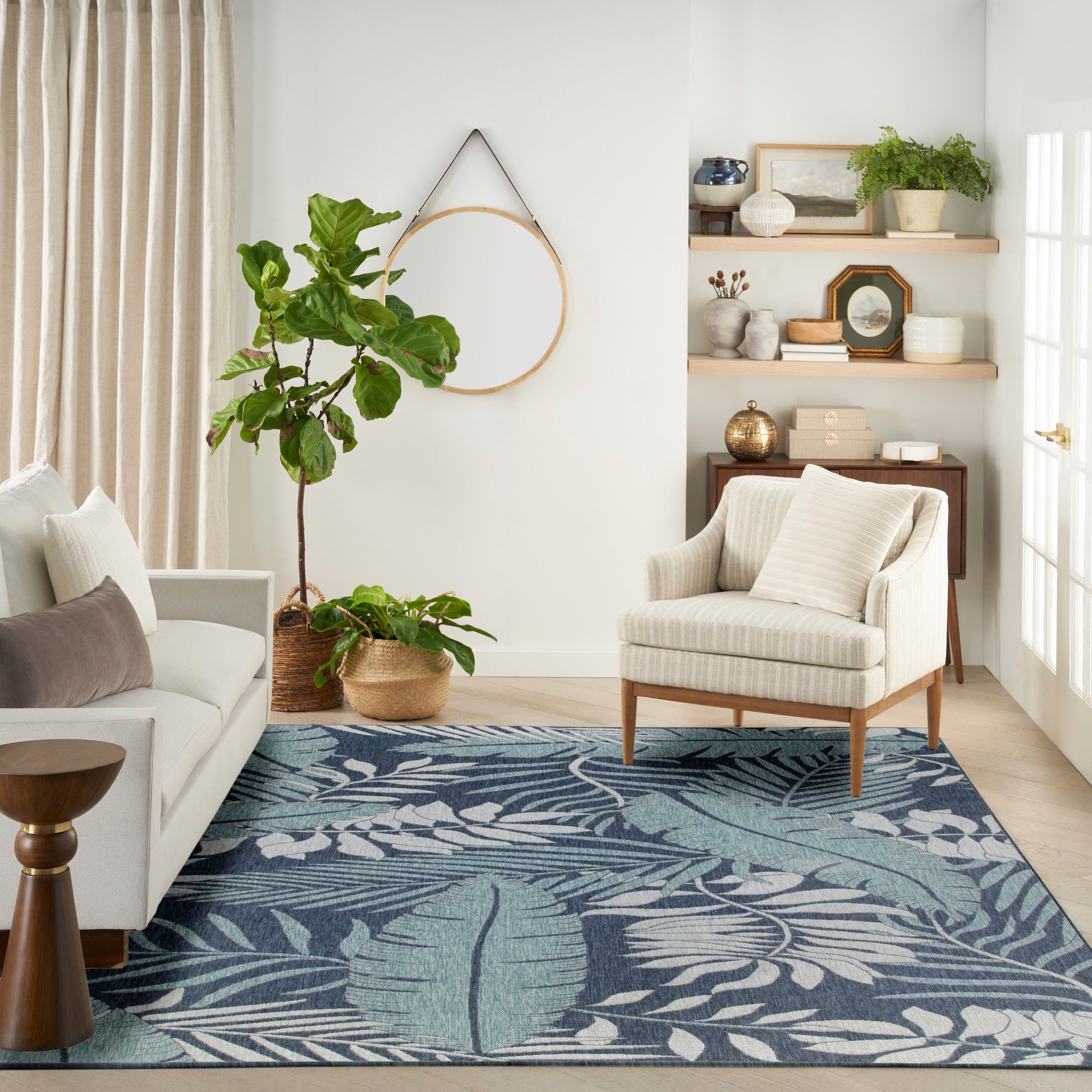 Cozy living room with beige striped armchair, grey sofa, blue botanical rug, tall potted plant, and wood shelves.