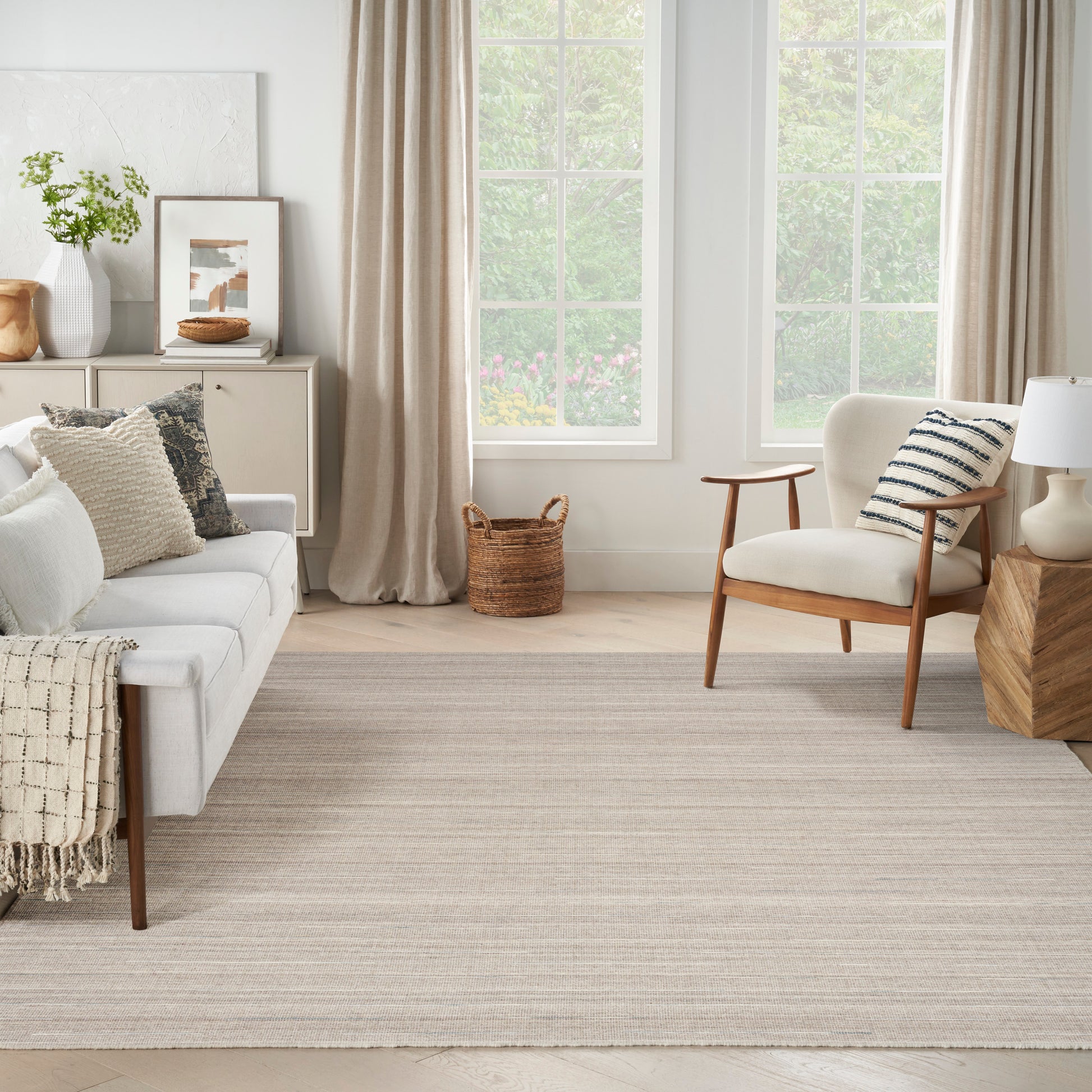 Bright living room with cream sofa, soft cushions, striped pillow, woven rug, wooden chair, wicker basket by large windows.