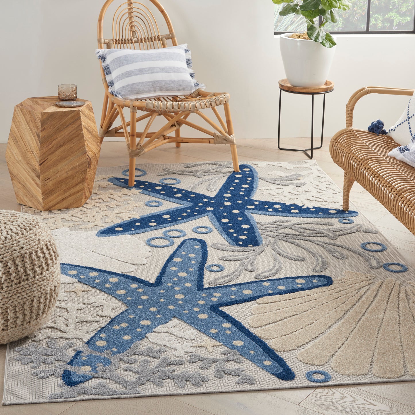 Coastal living room with blue starfish rug, rattan chairs, woven ottoman, striped cushions, and potted plant.