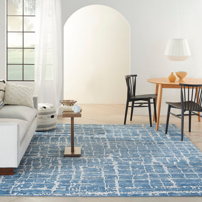 Bright airy living space with blue woven rug, light gray sofa, wooden side table, round dining table, black chairs, white pendant lamp, arched doorway.