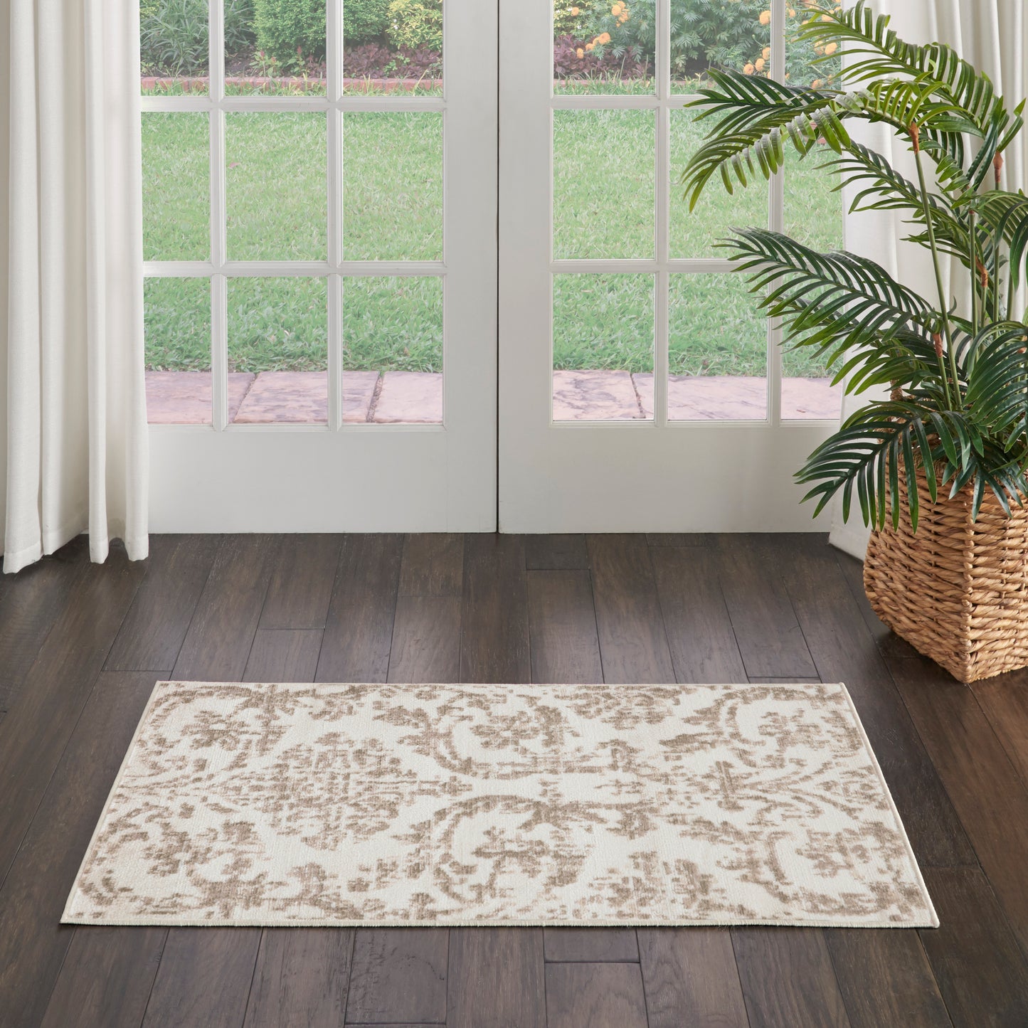 Rectangular beige damask-pattern area rug on dark wood floor beside glass French doors and a potted palm.