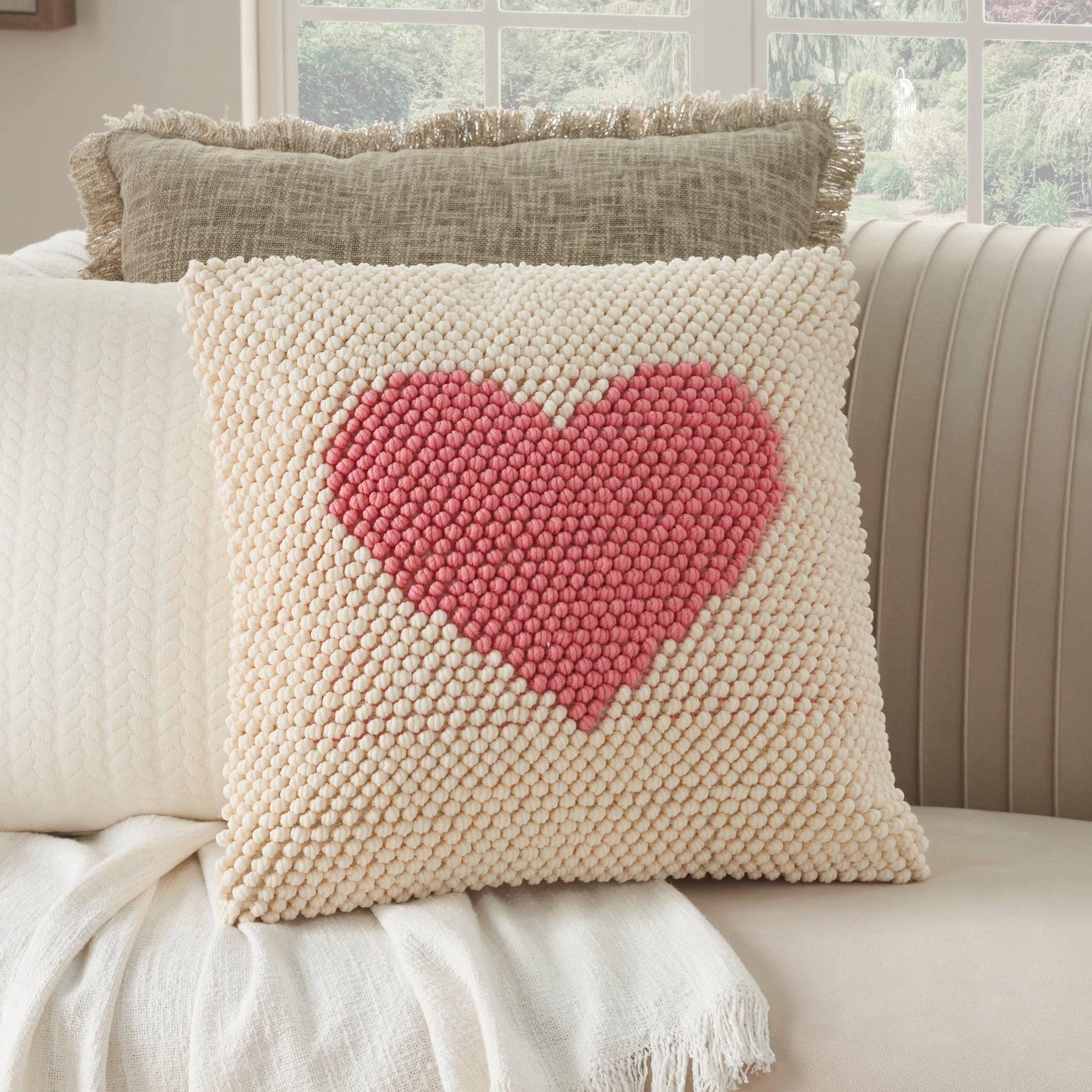 Cream textured pom-pom decorative pillow with a pink heart centerpiece on a beige sofa.