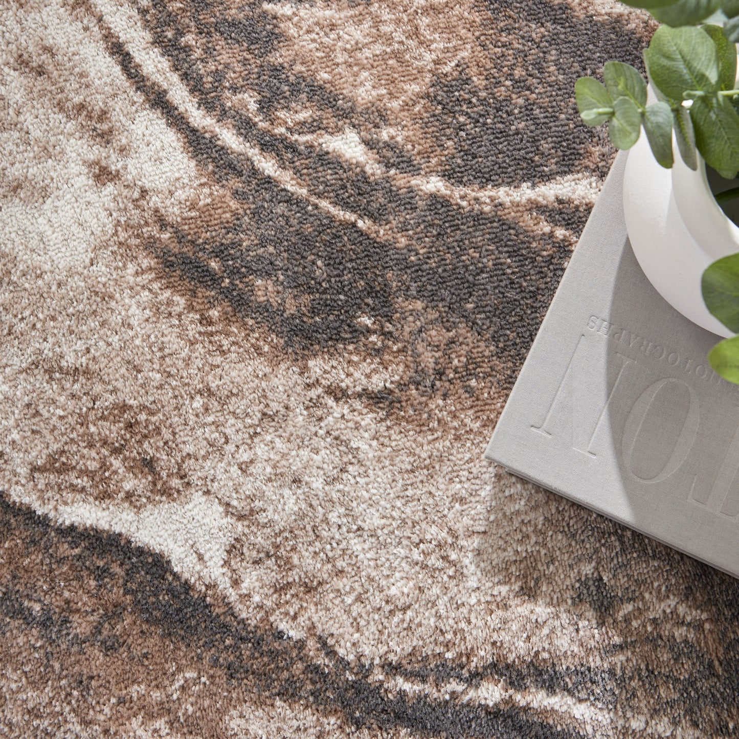 Earth-toned marble-pattern rug in beige and brown with a light gray decorative block and a white plant pot with green leaves.