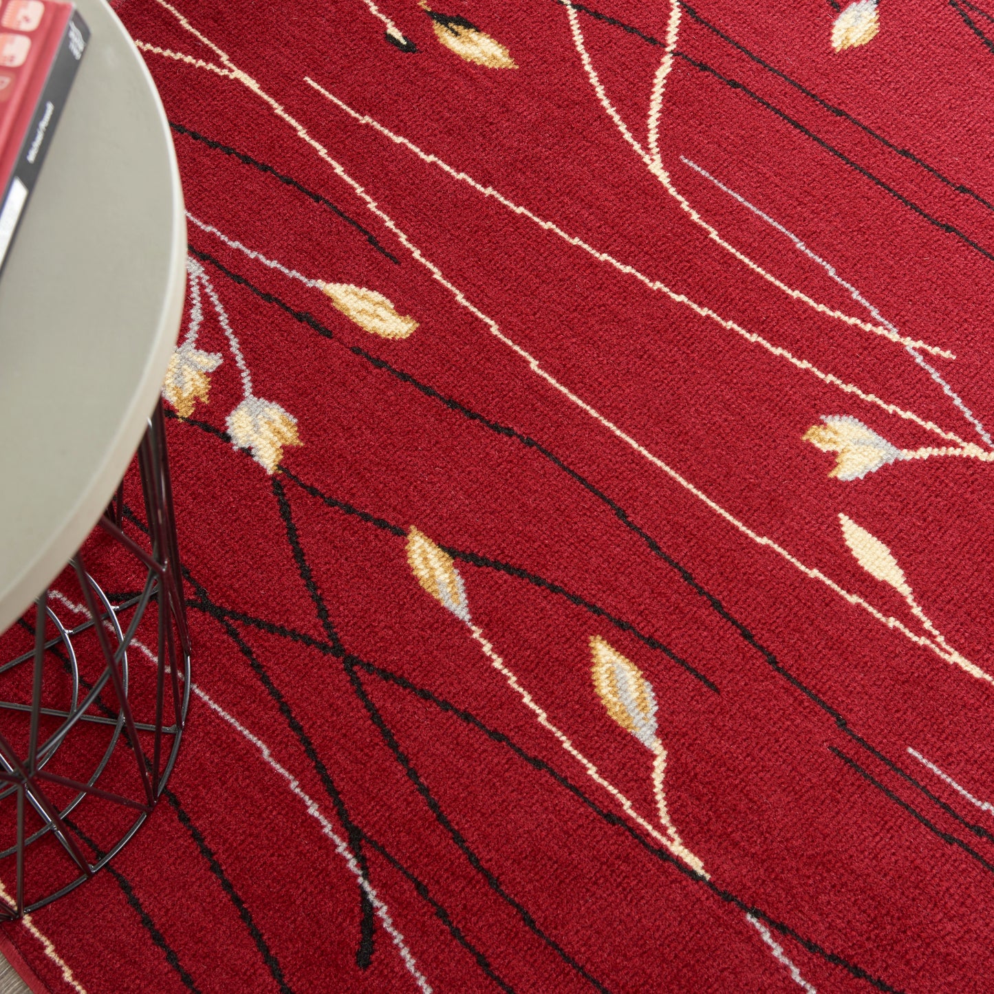 Red textured area rug with cream leaf motifs and black lines; modern wire-framed side table visible.