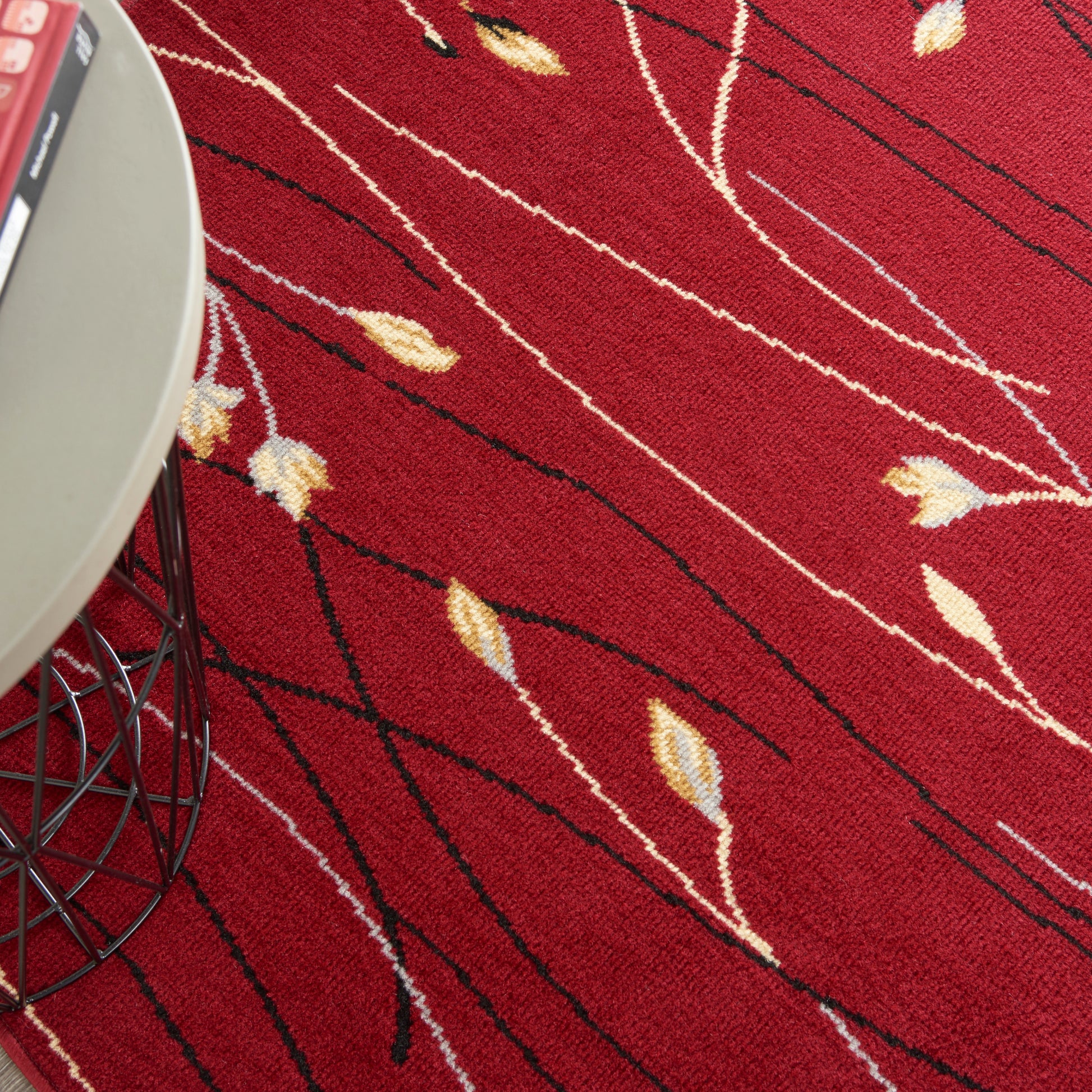 Red textured area rug with cream leaf motifs and black lines; modern wire-framed side table visible.
