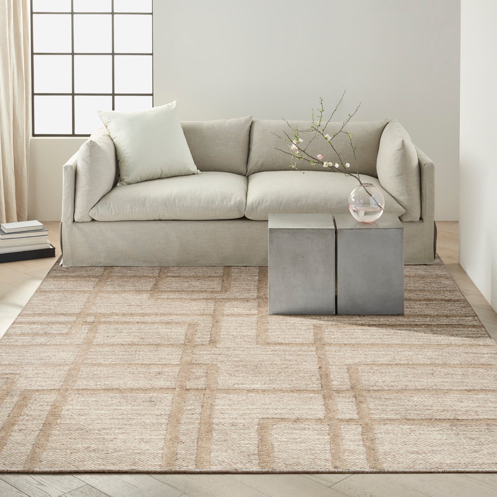 Neutral living room with light gray sofa, beige geometric rug, and gray block coffee table with a glass vase and branches.