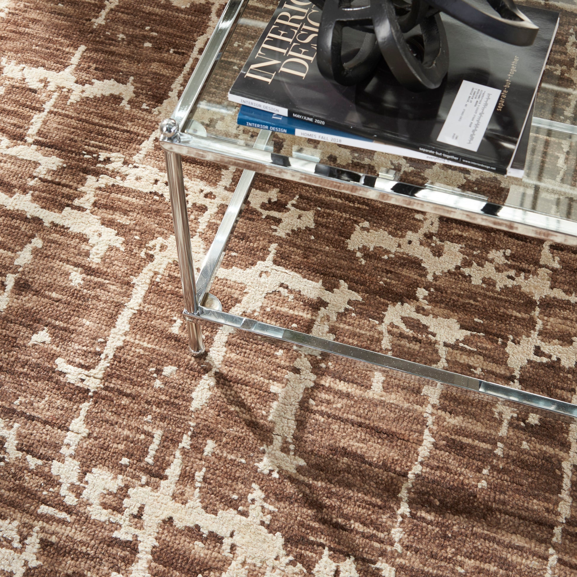 Rectangular glass coffee table with chrome frame on a brown patterned rug, magazines and a black sculptural decor piece on top.