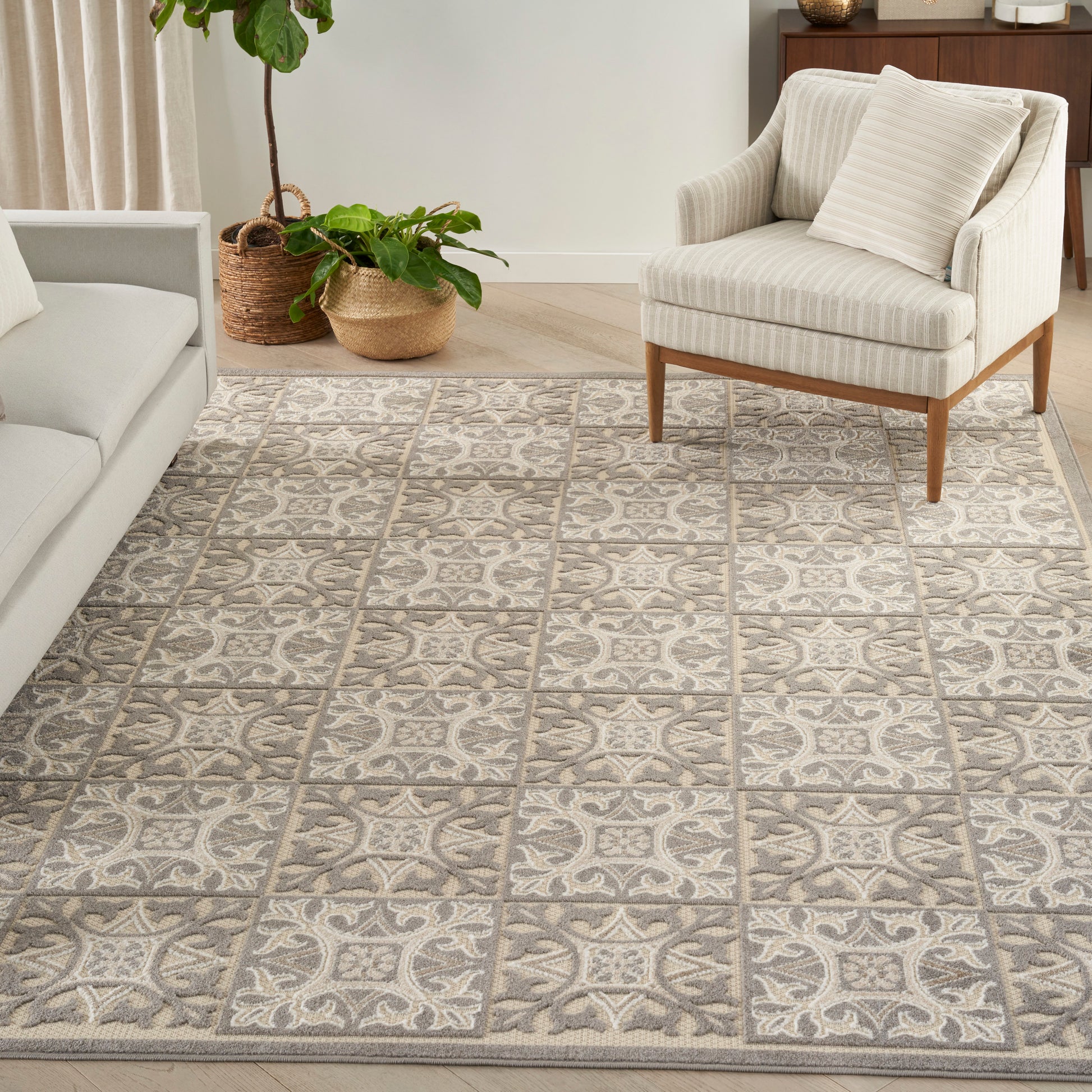 Neutral woven area rug with gray & cream square tile motif in a living room with beige sofa, striped armchair & plants.