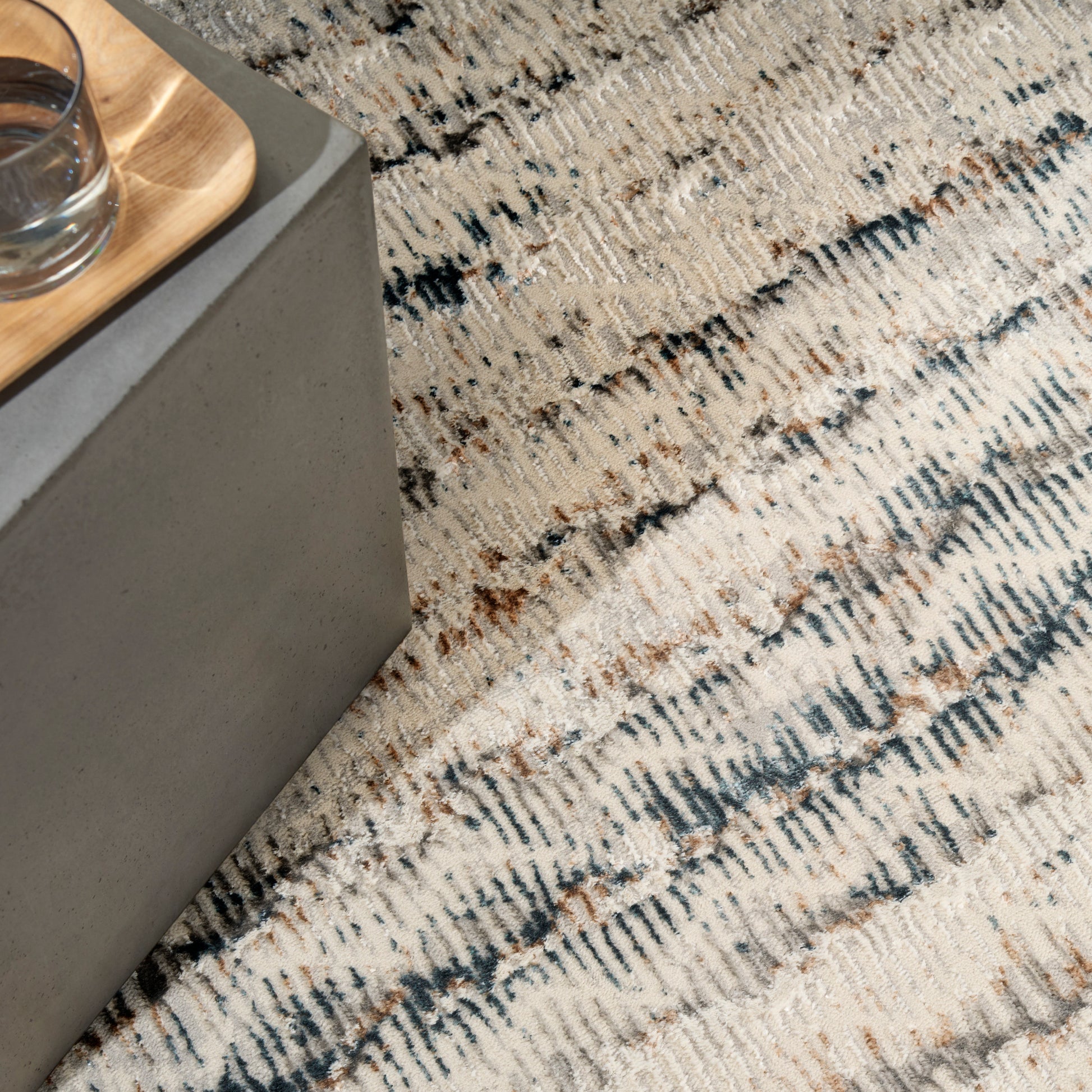 Grey concrete side table with a light wood tray and glass of water on a textured beige patterned rug.
