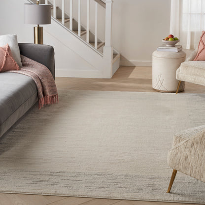 Modern living room with light gray sofa, blush throw, cream rug, white stairs, gold lamp, fruit bowl, woven ottoman.
