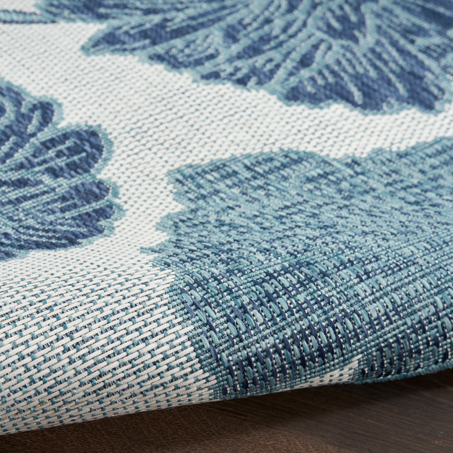 Close-up of a blue woven rug with teal accents, leaf pattern, folded edge on a wooden floor.