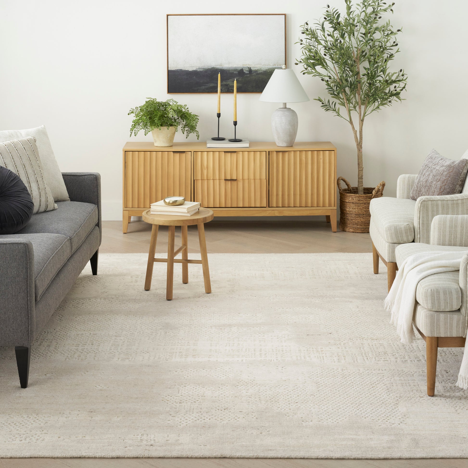 Neutral living room featuring a light-toned rug under a gray sofa and beige armchair, with a light wood slatted sideboard, greenery, candles, lamp and a tall plant.