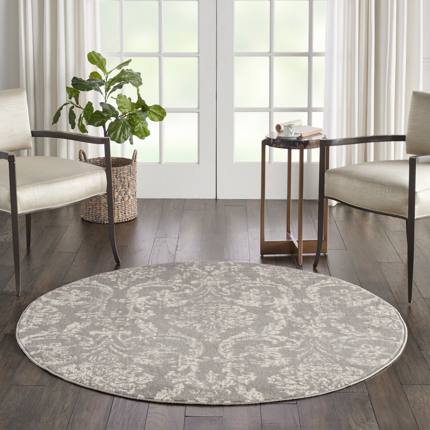 Round gray patterned area rug on dark wood floor between beige armchairs; plant in wicker basket left and small table near French doors.