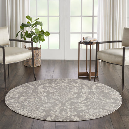 Round gray patterned area rug on dark wood floor between beige armchairs; plant in wicker basket left and small table near French doors.