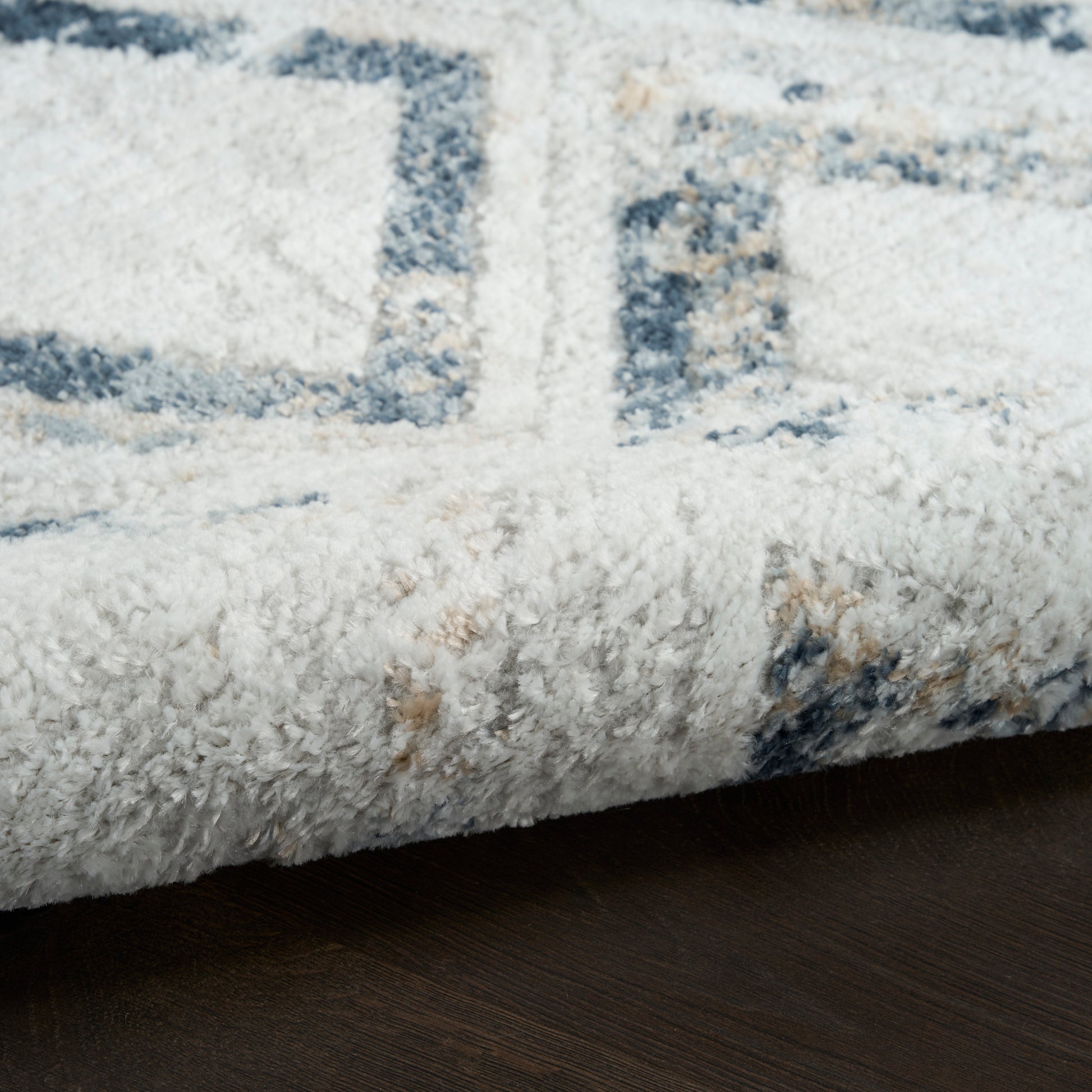 Close-up of a plush white rug with blue geometric pattern, folded edge resting on dark wood floor.