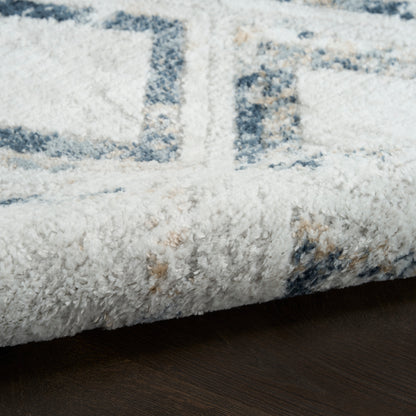 Close-up of a plush white rug with blue geometric pattern, folded edge resting on dark wood floor.