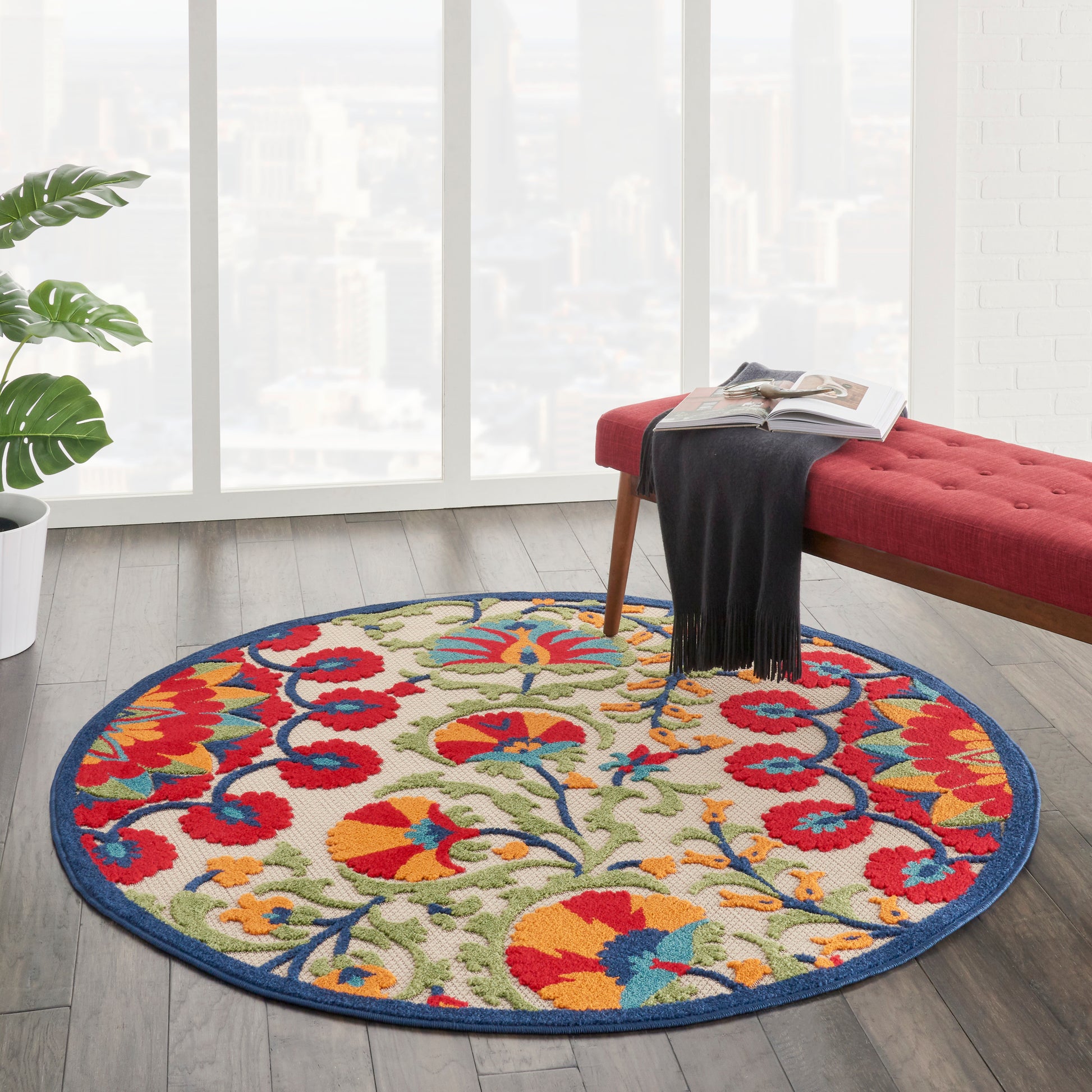 Round multicolor floral area rug with a blue border on a wood floor in a bright living room beside a red upholstered bench.