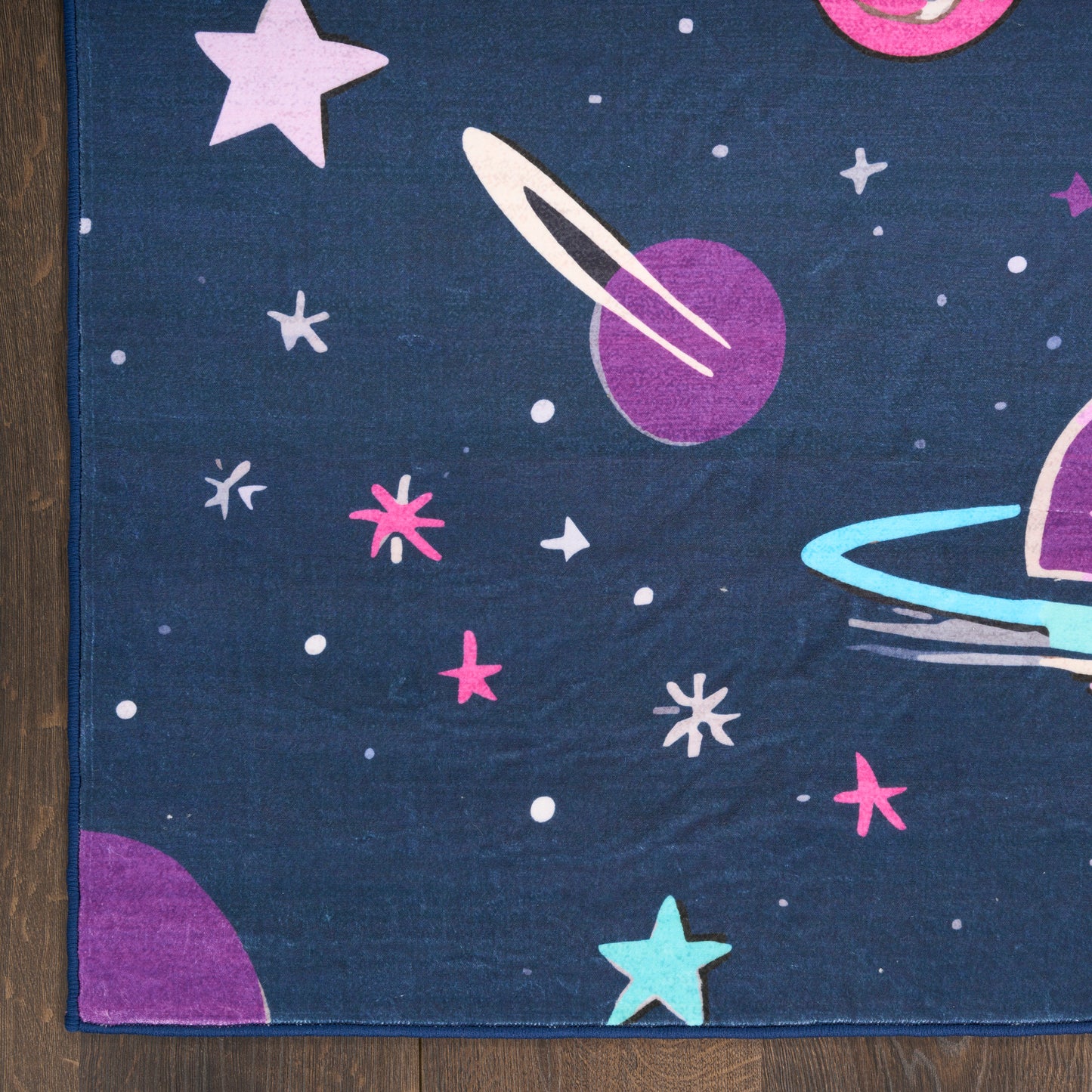 Dark blue space-pattern rug with purple planet and rings, turquoise ring, pink stars and white dots; wood floor edge visible.