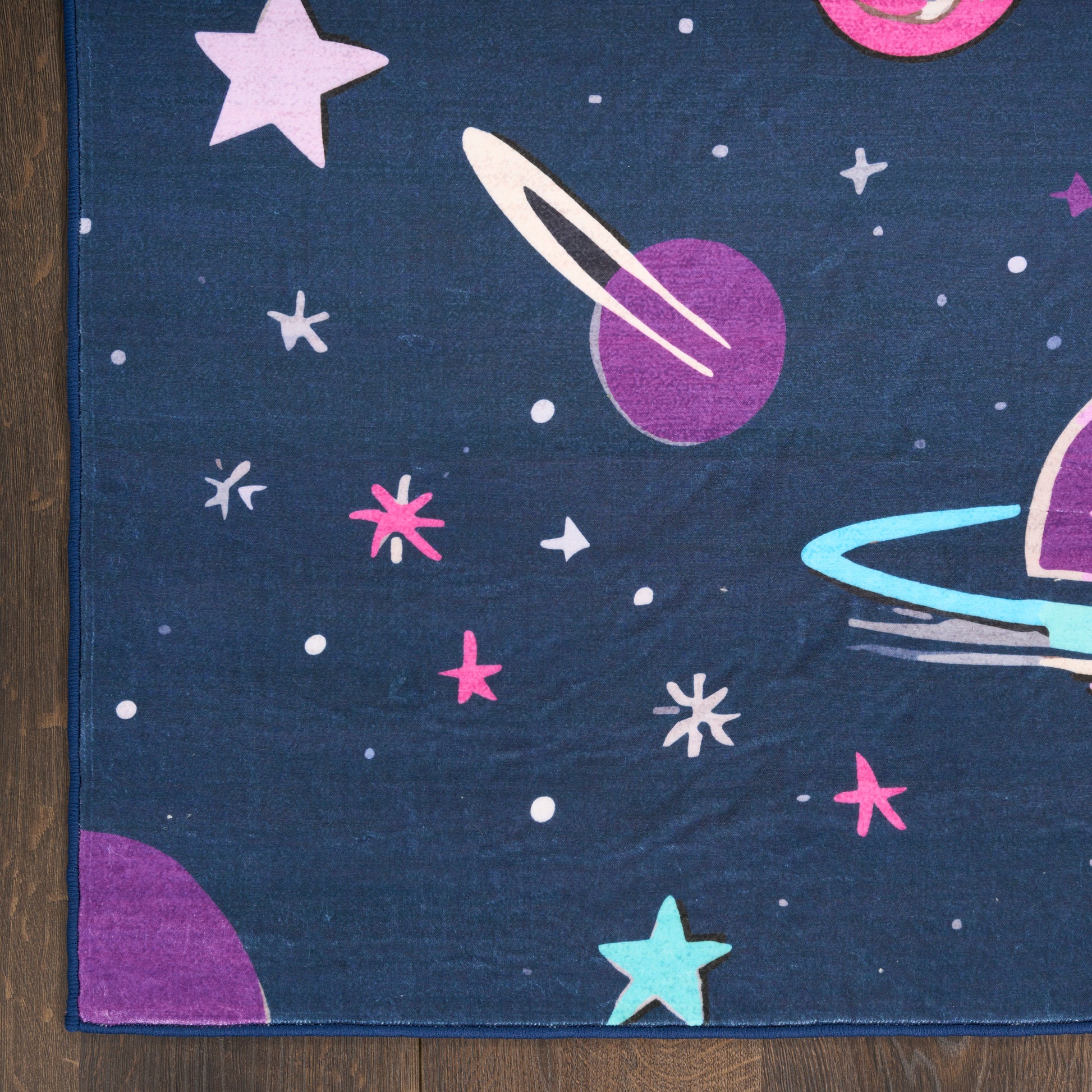 Dark blue space-pattern rug with purple planet and rings, turquoise ring, pink stars and white dots; wood floor edge visible.