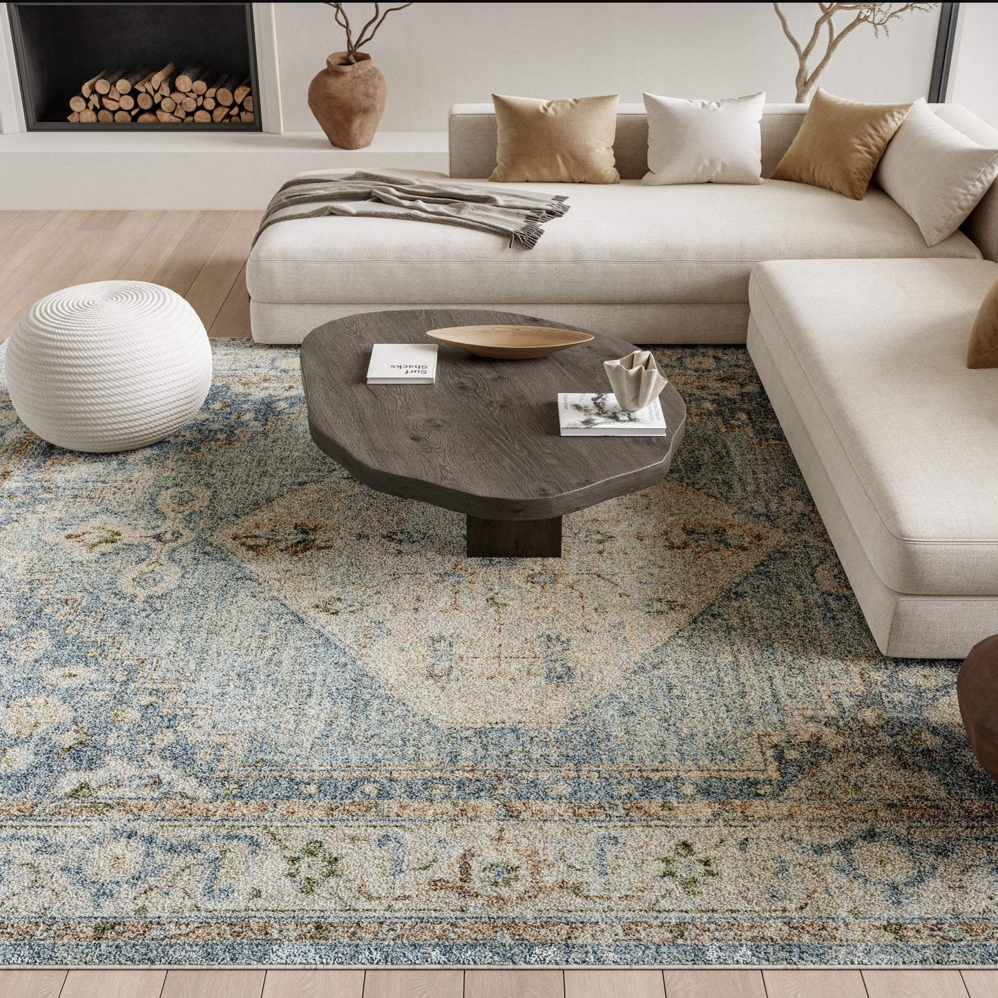 Cozy living room with beige sectional, dark wood coffee table, white knitted pouf, & blue-beige patterned rug