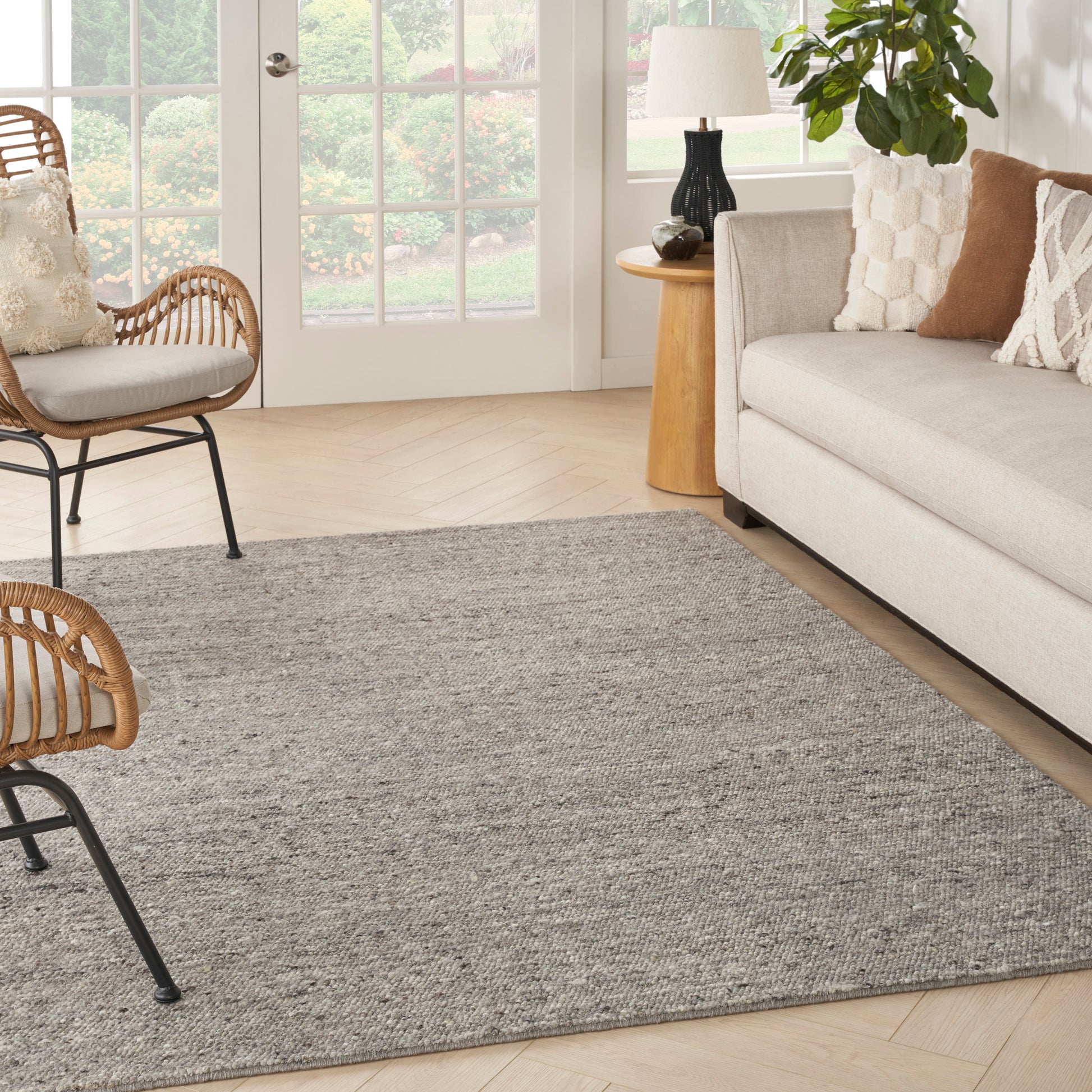 Soft gray textured area rug anchors a bright living room with beige sofa, rattan chairs, wooden side table, and greenery.
