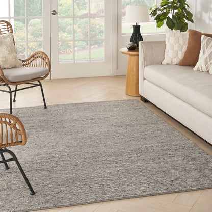 Soft gray textured area rug anchors a bright living room with beige sofa, rattan chairs, wooden side table, and greenery.