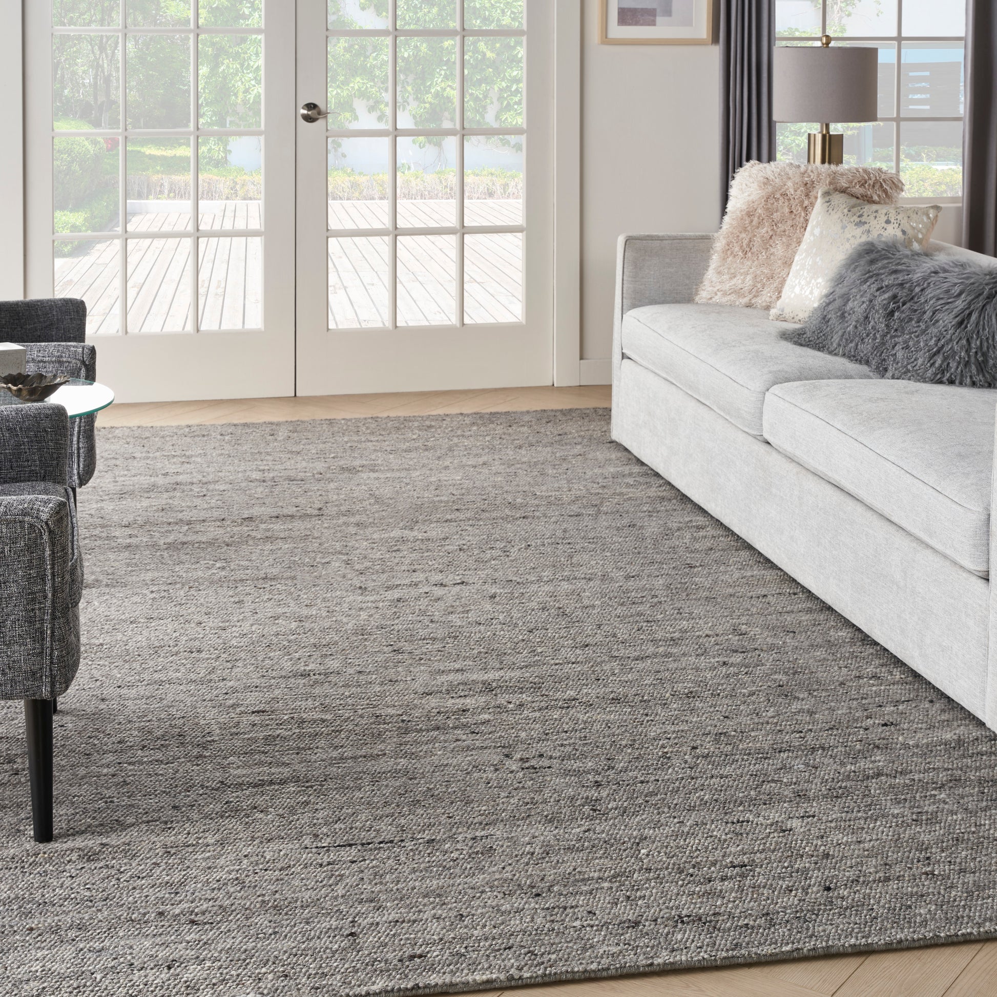 Living room with grey sofa, rug, French doors, beige and grey cushions, and a grey armchair with a glass side table.