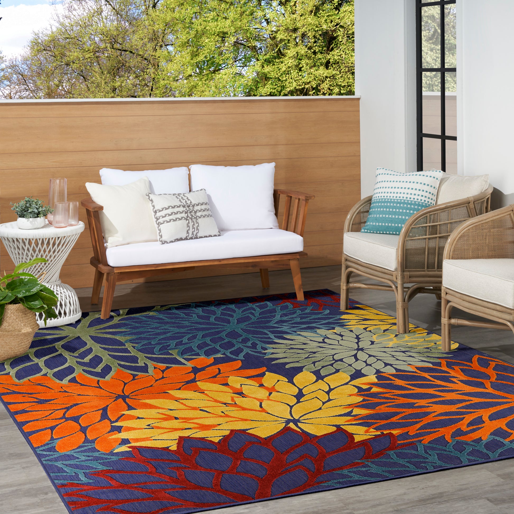 Outdoor patio with leaf-pattern rug in orange, yellow, blue; white-cushioned wooden sofa, wicker chairs, potted plant.