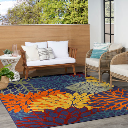 Outdoor patio with leaf-pattern rug in orange, yellow, blue; white-cushioned wooden sofa, wicker chairs, potted plant.