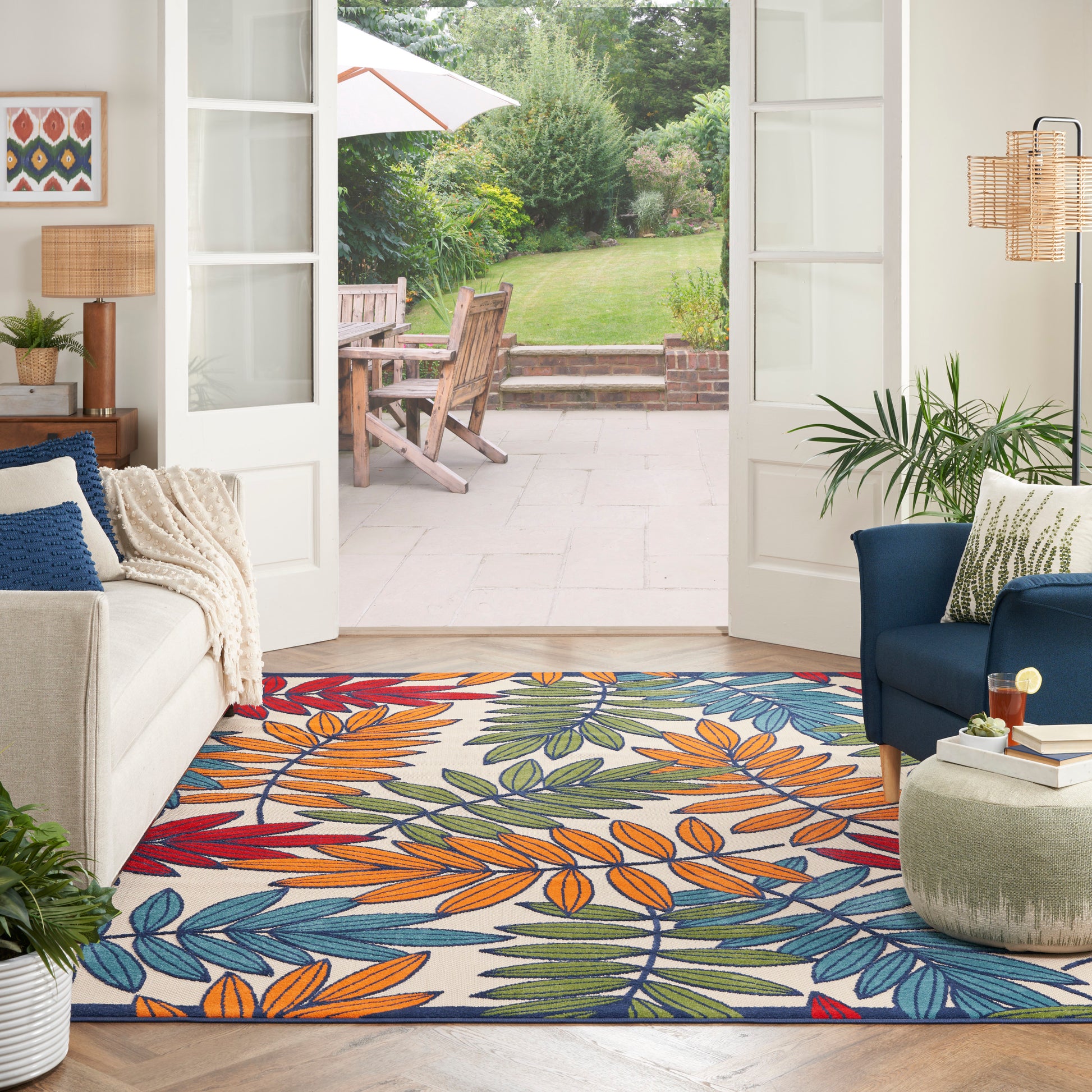Bright living room with French doors to garden; colorful tropical leaf rug in orange, blue & green; beige fabric sofa, blue chair.