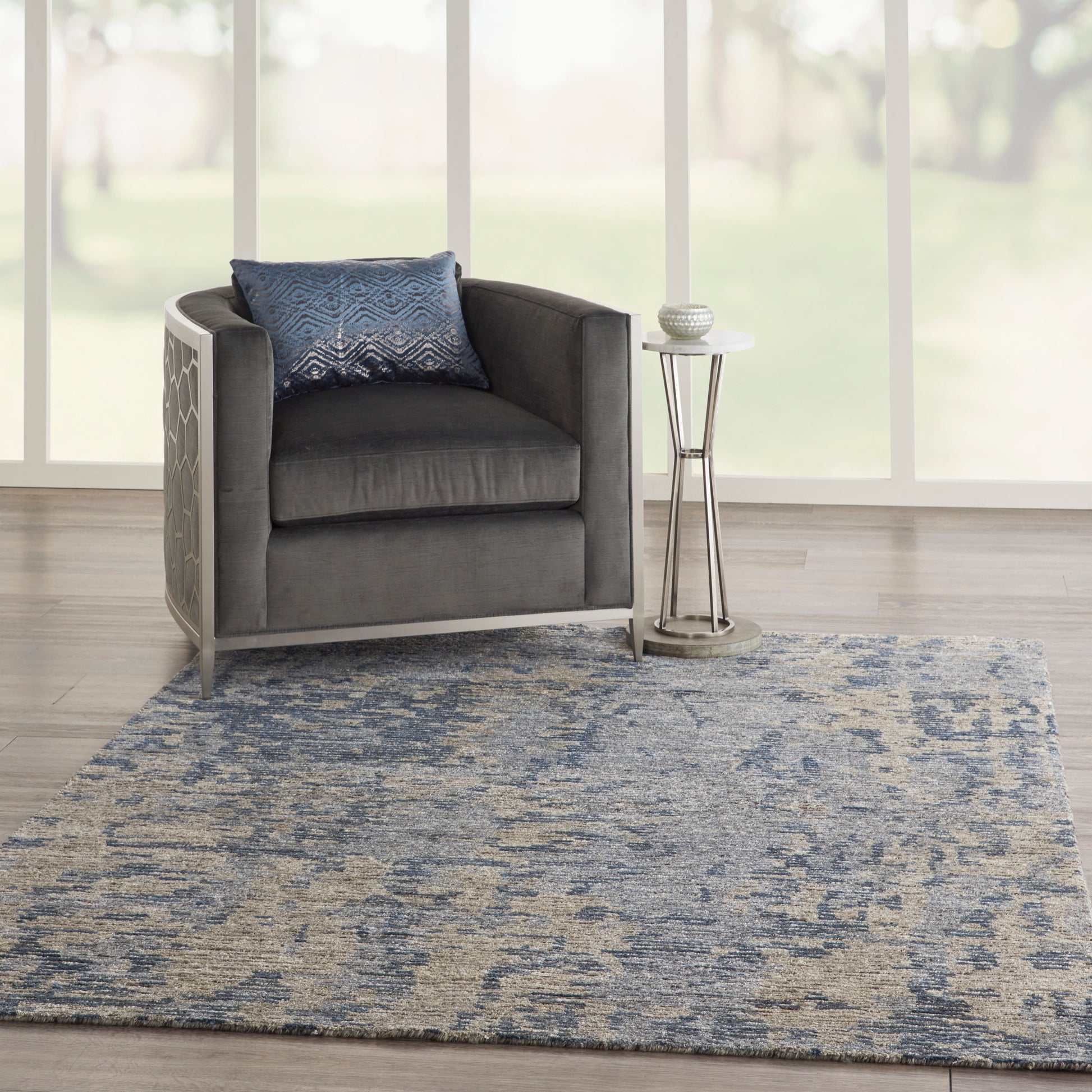 Gray armchair with metal frame, blue patterned pillow, chrome side table, and blue-gray abstract rug in a bright living room.