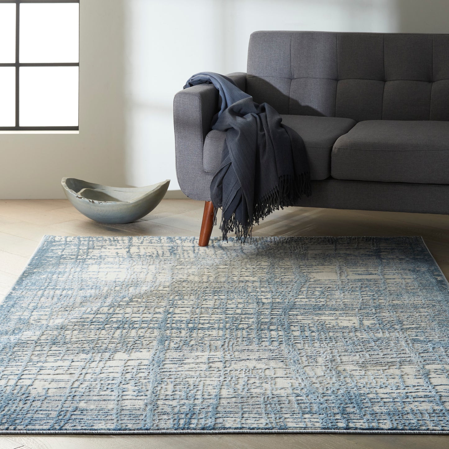 Gray fabric sofa with wooden legs, navy throw; blue geometric rug on light wood floor, ceramic bowl nearby.