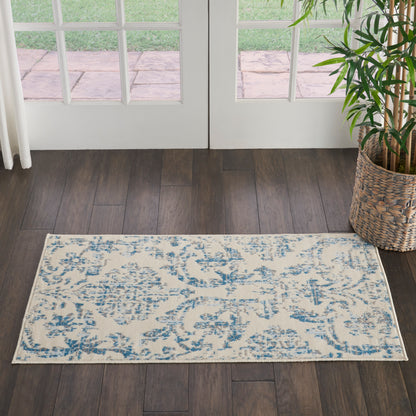 Rectangular cream rug with blue floral pattern on dark wood floor by white French doors; plant in woven basket nearby.