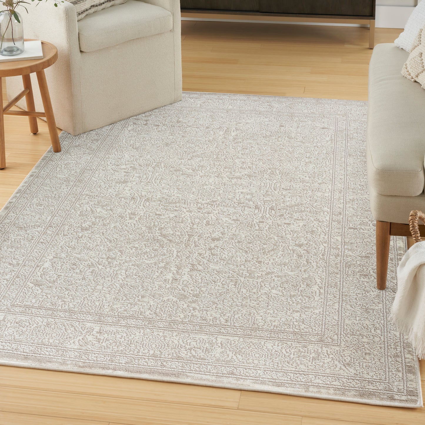 Light gray patterned area rug with a delicate border, laid over a wooden floor in a cozy living room with beige sofas.