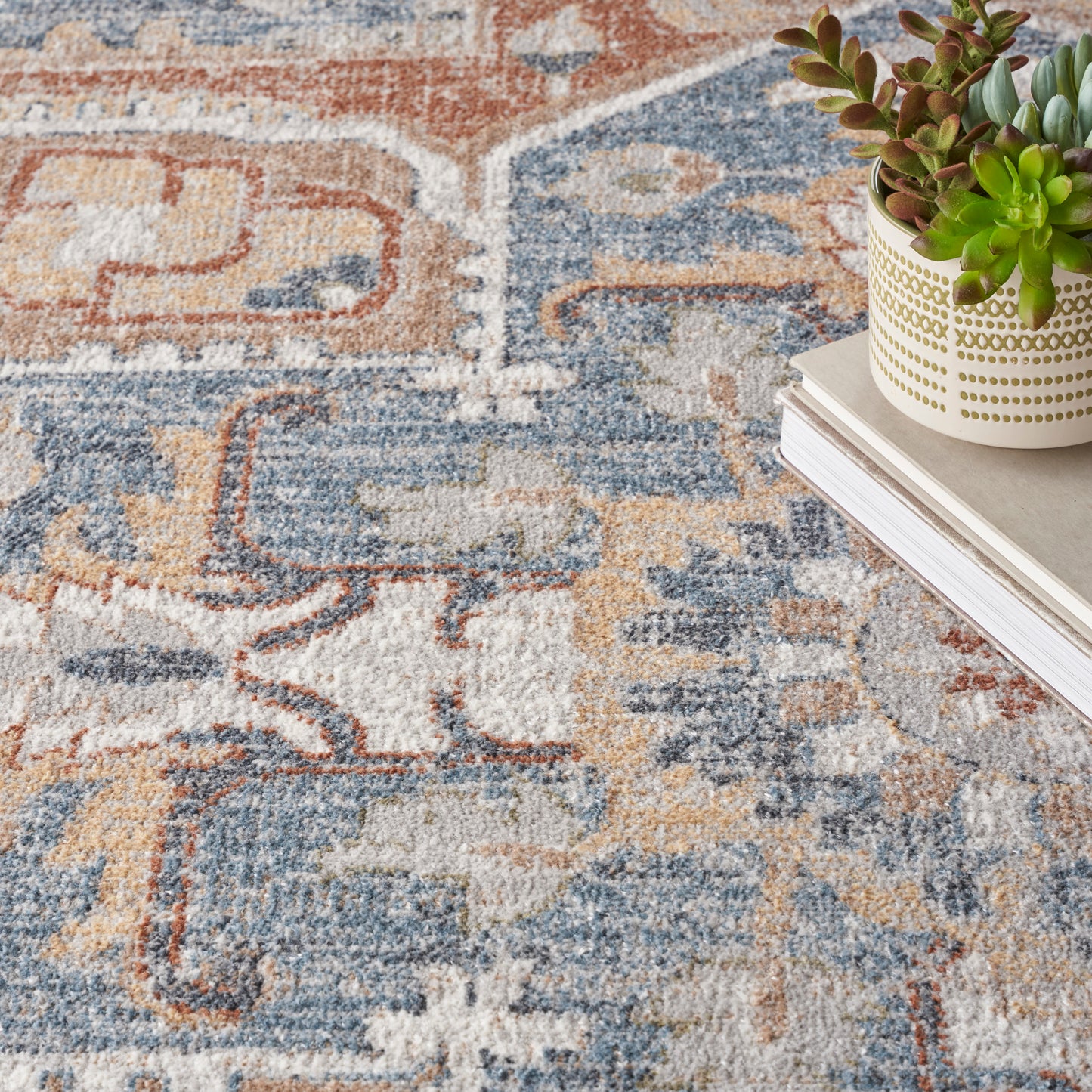 Close-up of an oriental rug in blue, tan and orange; white pot with gold dotted pattern and succulents atop stacked books.