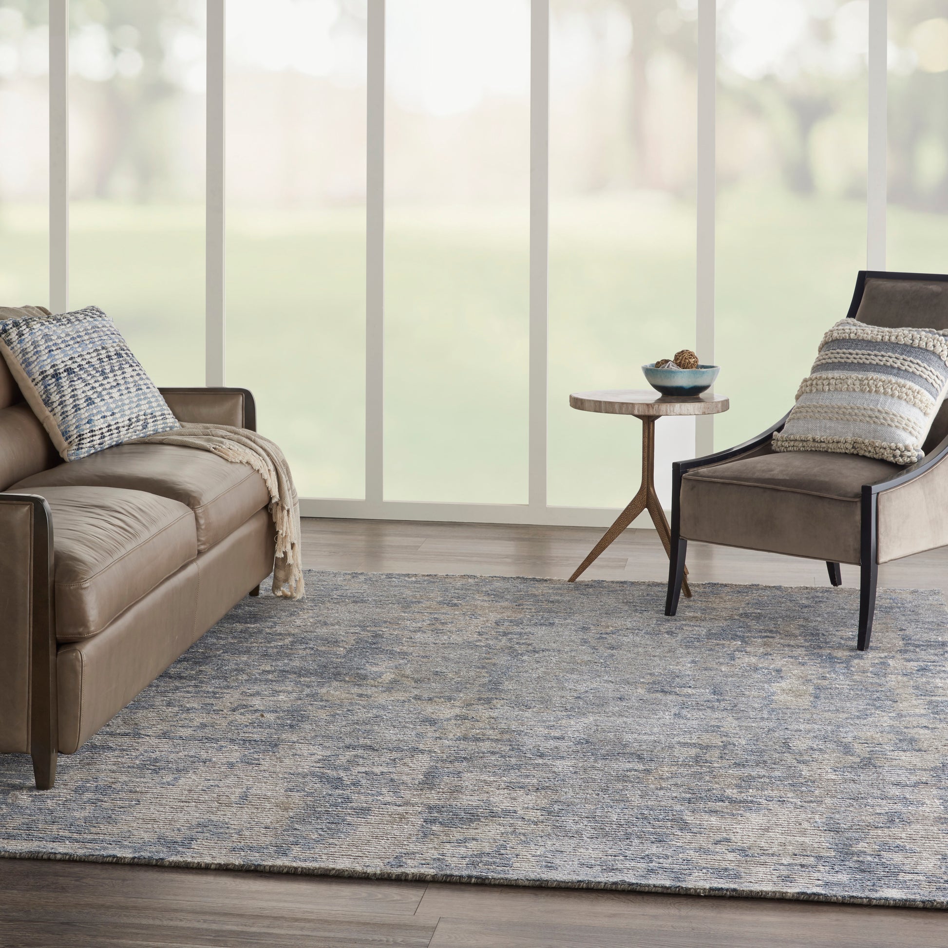 Brown leather sofa and matching chair, blue-gray area rug, neutral throws, small round side table under large windows.