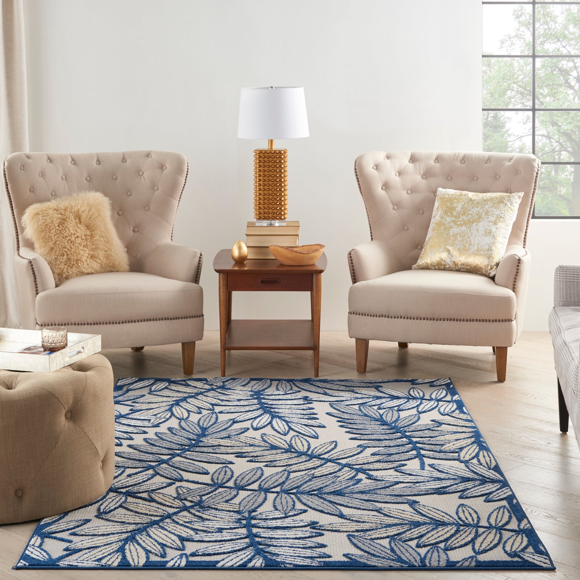 Two beige tufted armchairs with a tufted ottoman, wooden side table, gold lamp, and a blue botanical rug.