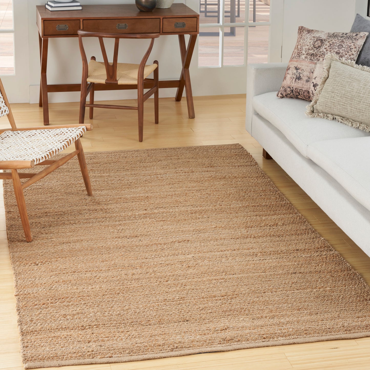 Natural jute rectangular area rug in a bright living room with a wooden desk, chair, woven coffee table and cream sofa.