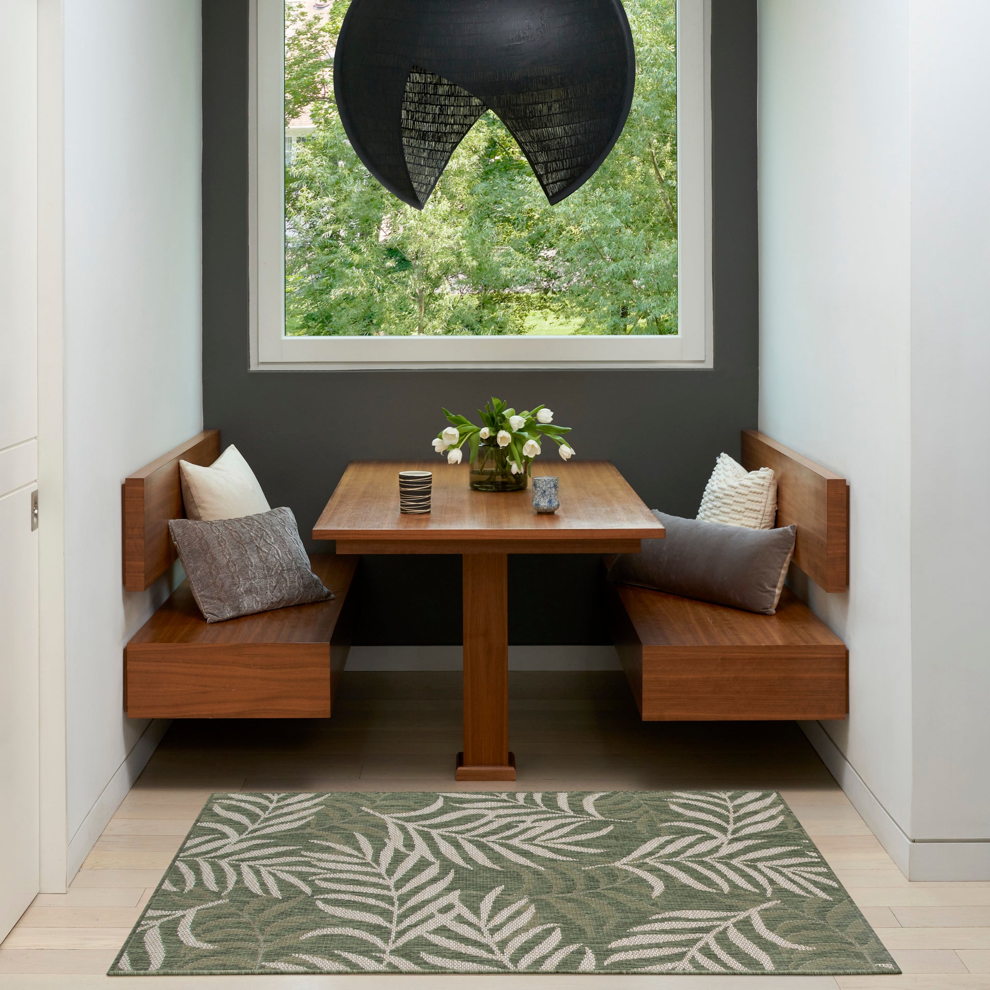Wood corner dining nook with square table, L-shaped benches, cushions, white flowers centerpiece, black pendant light, green rug.