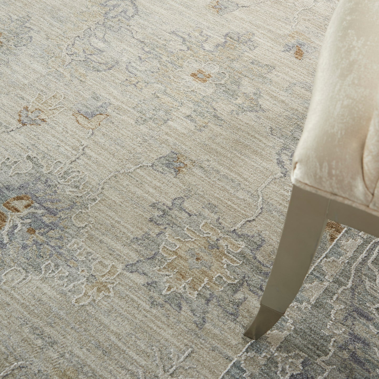 Soft beige-gray ornate patterned area rug with taupe accents beside a beige upholstered chair on a tapered metal leg.