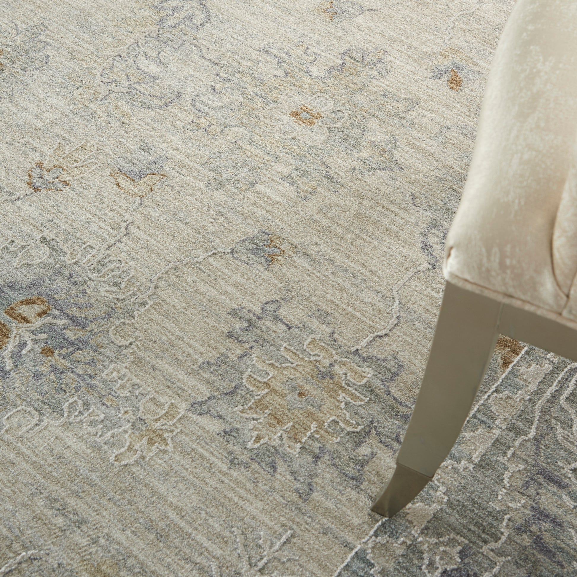 Soft beige-gray ornate patterned area rug with taupe accents beside a beige upholstered chair on a tapered metal leg.