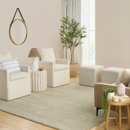 Cozy neutral living room with cream armchairs, round wooden mirror, striped side table, potted plant, and beige rug.