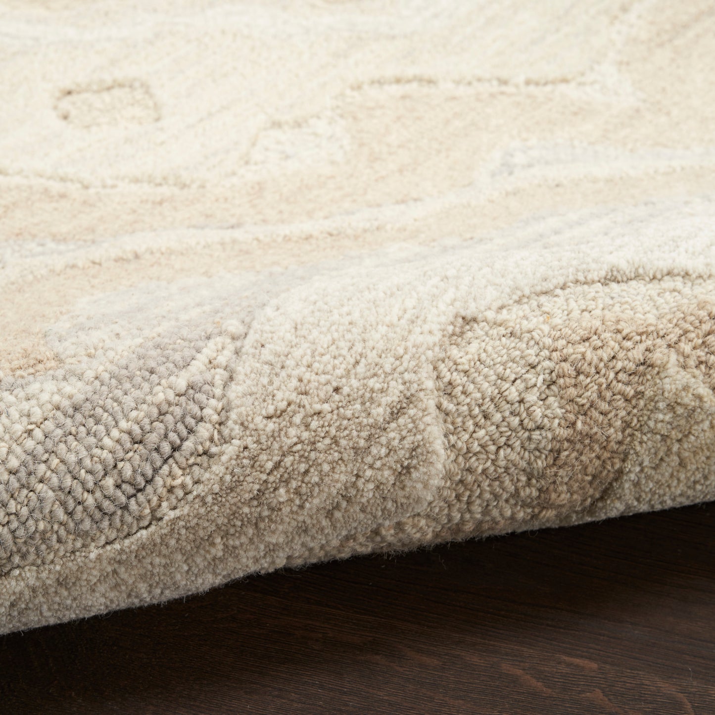 Beige and cream tufted plush rug with textured pile and subtle pattern, edge curled over on dark wood floor.