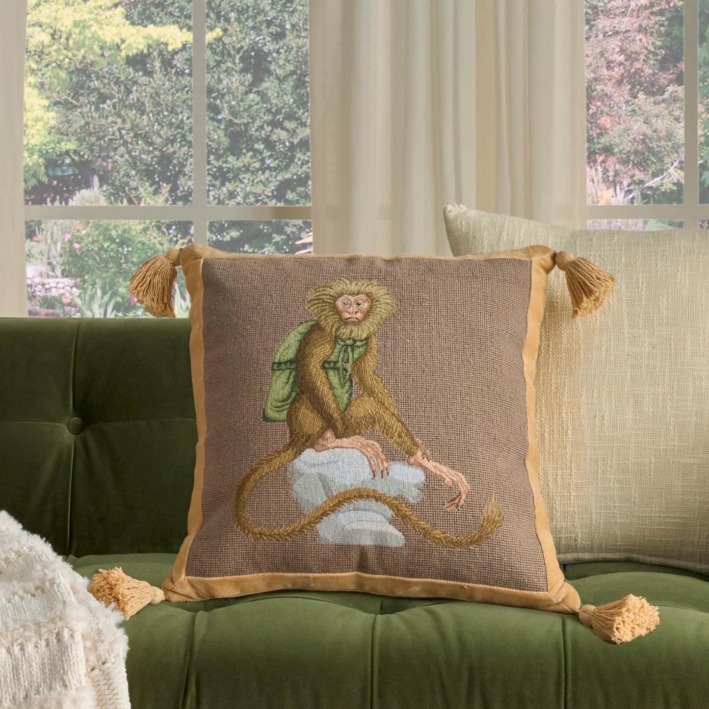 Decorative square pillow on a green tufted velvet sofa; brown burlap-like cover, tan border and tassels, with a stitched monkey in a green scarf.