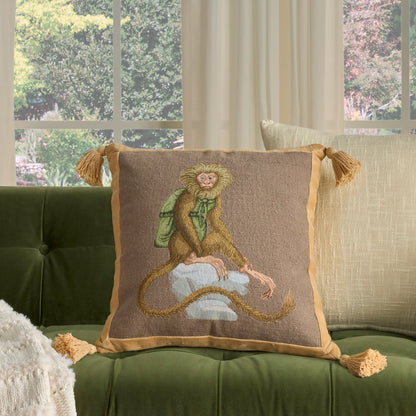 Decorative square pillow on a green tufted velvet sofa; brown burlap-like cover, tan border and tassels, with a stitched monkey in a green scarf.