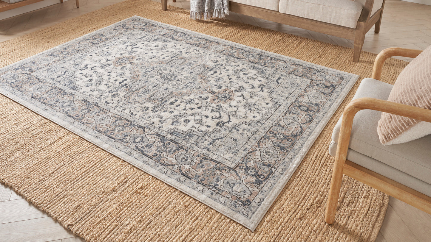 Light gray-blue oriental-style rug with floral border over natural jute runner in a cozy living room with wooden furniture.