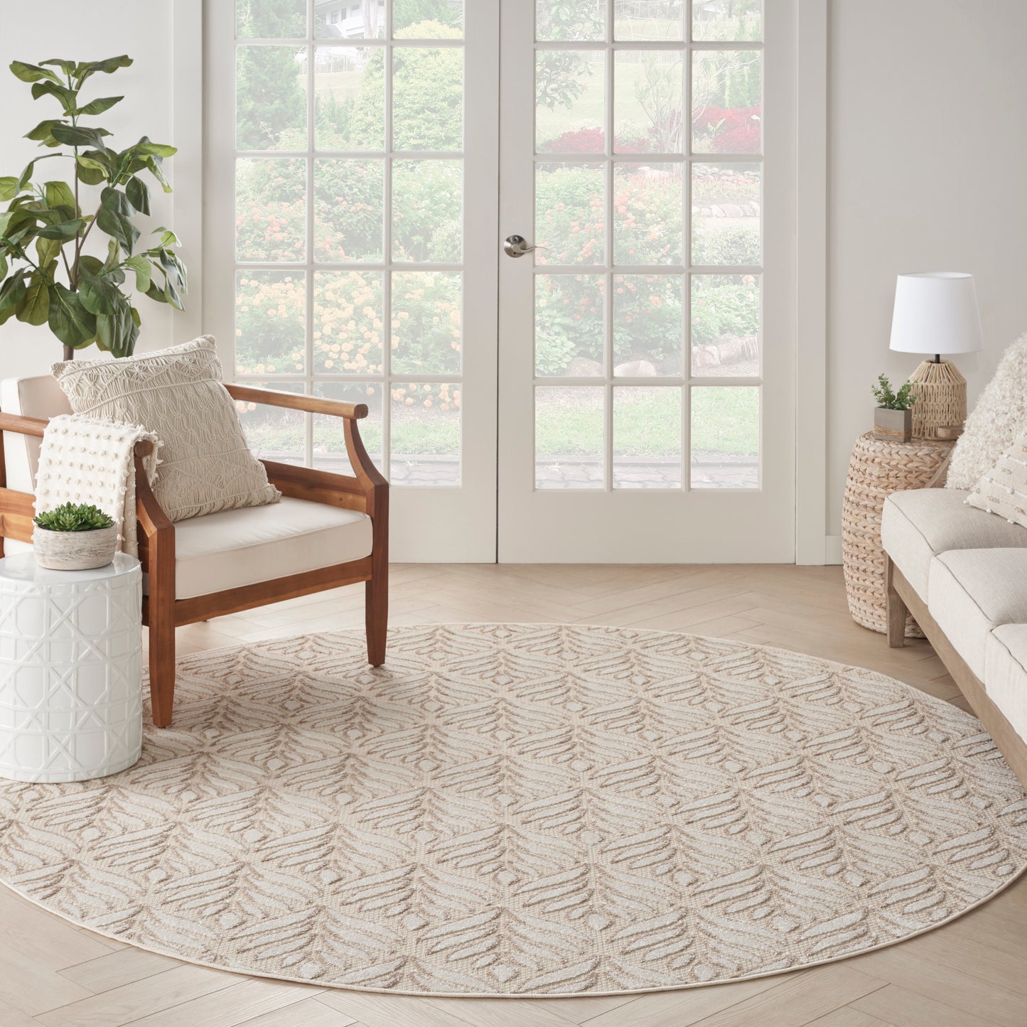 Round beige rug with subtle leaf pattern in a bright neutral living room; wooden armchair, woven side table, potted plants.