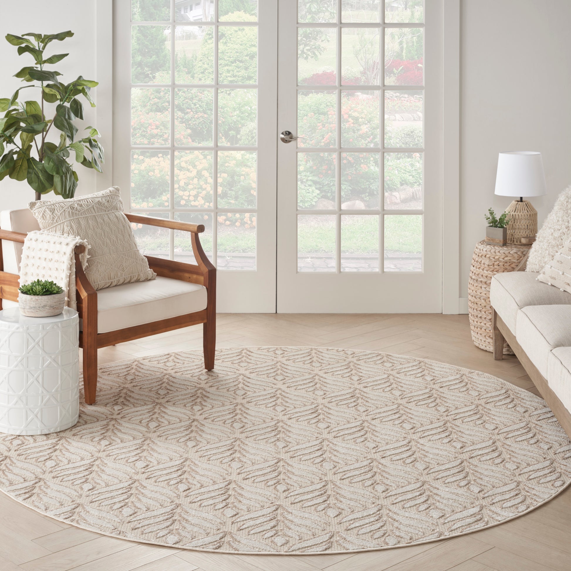 Round beige rug with subtle leaf pattern in a bright neutral living room; wooden armchair, woven side table, potted plants.